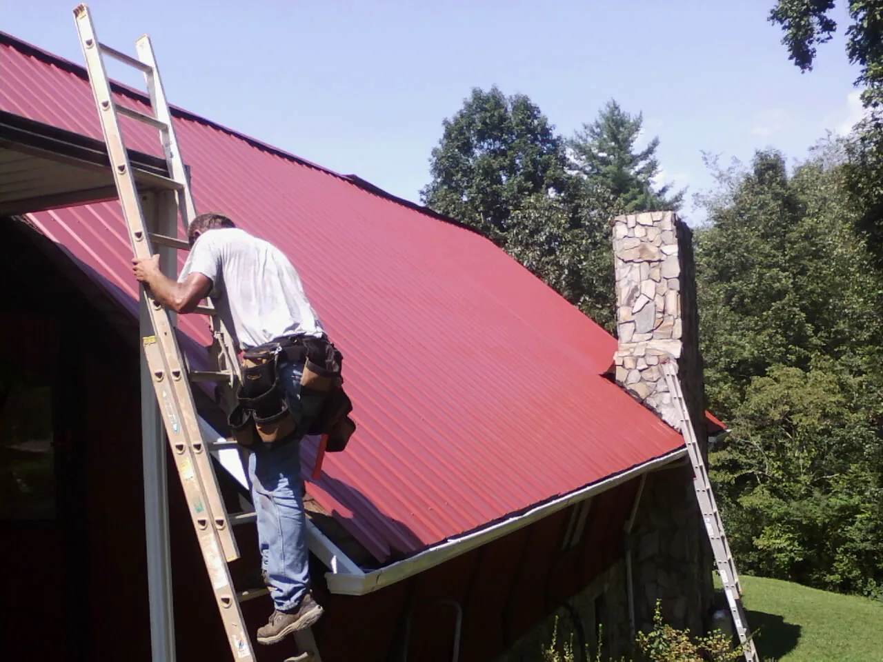 Roofer on a ladder beside a red metal roof and stone chimney — Soffit & Fascia Repair by Roofing Brothers in Dallastown